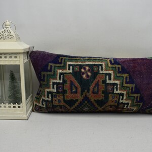 May include: A long rectangular cushion with a colourful geometric pattern in shades of purple, green, orange, and white. The cushion is made of a soft, woven fabric and has a fringed edge.