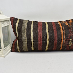 May include: A long, rectangular pillow with a striped pattern in shades of brown, red, orange, and cream. The pillow is made of woven fabric and has a slightly textured surface.