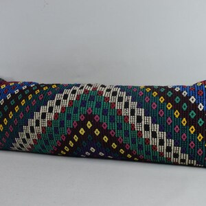 May include: A long, rectangular pillow with a colorful geometric pattern. The pattern features squares, diamonds, and dots in shades of blue, green, red, yellow, and black.