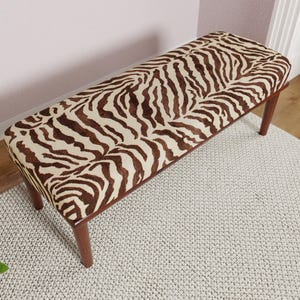 May include: A rectangular bench with a zebra print seat cushion and four wooden legs. The cushion features a brown and cream-colored zebra pattern. The bench is a decorative furniture piece.