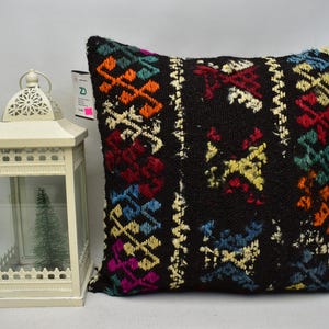 May include: A decorative black pillow with a colorful geometric pattern in orange, red, blue, and yellow. A white lantern with a small Christmas tree inside is next to the pillow.