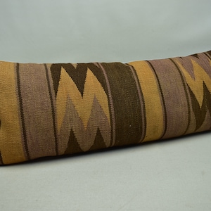 May include: A long, rectangular cushion with a brown, beige and purple geometric pattern. The cushion is made of woven fabric and has a fringed edge.