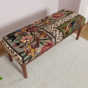 Puede incluir: Banco rectangular con un estampado floral en tonos rojos, amarillos y verdes. El banco tiene un fondo verde claro con un dise&ntilde;o floral detallado. Las patas son de madera oscura.