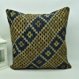 May include: A square cushion with a geometric pattern in shades of blue, yellow, and brown. The cushion is made of a woven fabric and has a fringe border.