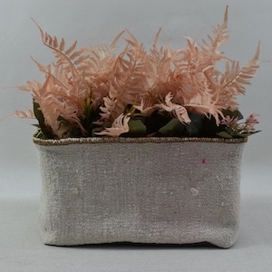 May include: A white fabric planter with a brown trim, filled with pink artificial ferns.