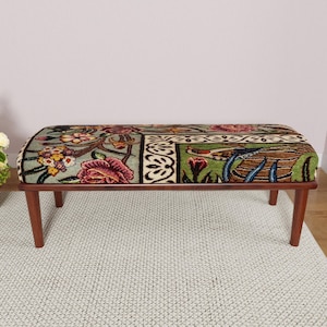 Puede incluir: Banco rectangular con un coj&iacute;n estampado floral en tonos verdes, rosas y marrones. El banco tiene un marco de madera marr&oacute;n oscuro y cuatro patas anguladas. El coj&iacute;n presenta un dise&ntilde;o bot&aacute;nico detallado.