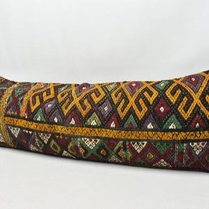May include: A long, rectangular pillow with a detailed, geometric pattern in shades of gold, burgundy, green, and black. The pillow has a textured appearance and a decorative border.