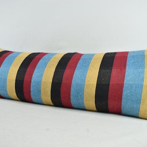 May include: A long, rectangular pillow with a striped pattern. The stripes alternate between blue, yellow, black, and red. The pillow is placed on a white surface.