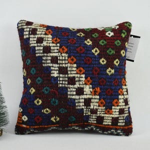 May include: A square cushion with a colourful geometric pattern in shades of red, blue, green, and white. The cushion is made of woven fabric and has a fringed edge.