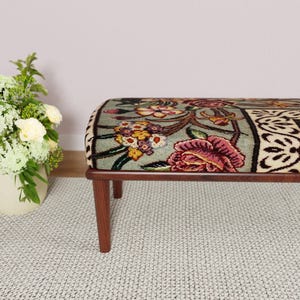 Puede incluir: Un banco con estampado floral y marco de madera. El banco presenta una parte superior rectangular con un dise&ntilde;o colorido de flores en tonos rosa, amarillo y verde. Las patas son de color marr&oacute;n oscuro.