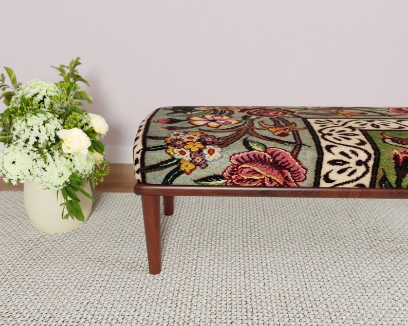 Puede incluir: Un banco con estampado floral y marco de madera. El banco presenta una parte superior rectangular con un dise&ntilde;o colorido de flores en tonos rosa, amarillo y verde. Las patas son de color marr&oacute;n oscuro.