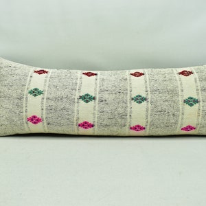 May include: A long, rectangular cushion with a white background and a grey and white striped pattern. The cushion has a geometric design with pink, green and red accents.