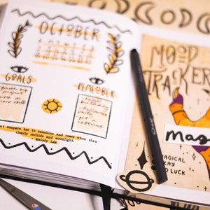 Puede incluir: Una libreta blanca con un diseño amarillo y negro. La libreta tiene un calendario para octubre, una sección para objetivos y una sección para recordatorios. La libreta también tiene una cita sobre comparar a alguien con el sol y las rosas. La libreta está abierta en una página con un rastreador de humor que tiene una mano con un diseño morado y naranja y la palabra "magia" escrita en ella.