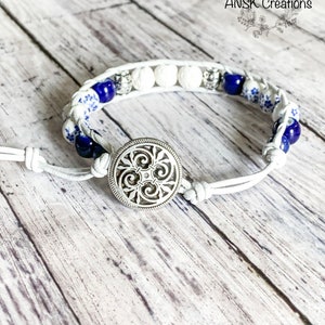 May include: White leather bracelet with blue and white ceramic beads, white lava beads, and a silver metal button closure. The bracelet has a knotted design and the text "ANSK Creations" is visible in the background.