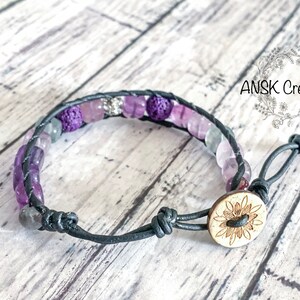 May include: A purple and green gemstone bracelet with a black leather cord and a wooden button closure. The button has a carved sunflower design. The bracelet is on a wooden surface.