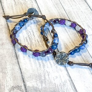 May include: Two brown leather bracelets with purple, blue, and white beads. One bracelet has a silver flower charm.