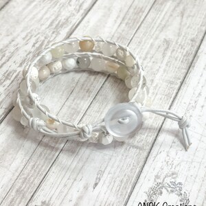 May include: A white wrap bracelet with cream-colored and white beads. The bracelet is made of white leather and has a large white button closure. The bracelet is designed to wrap around the wrist multiple times.
