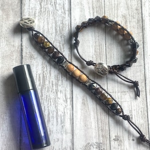 May include: Two beaded bracelets with brown, tan, and black stones, and a blue essential oil bottle with a black cap. The bracelets are on a wooden surface. The text "ANSK Creations" is visible.