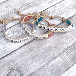 May include: Three handmade bracelets with the words "MAMA BEAR" spelled out in white letter beads. The bracelets are made with a braided leather band and jute cord. Each bracelet has a different color bead accent: purple, pink, and blue. The bracelets are on a light wood surface.