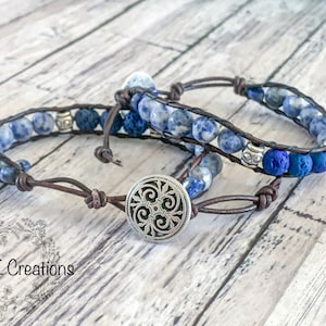 May include: Two leather bracelets with blue and white gemstone beads and dark blue lava beads. One bracelet has a silver charm with a decorative design. The bracelets are on a wooden surface.