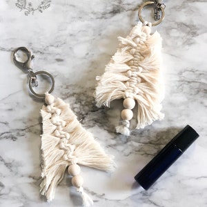 May include: Two white macrame Christmas tree keychains with wooden beads and silver key rings. The keychains are on a white marble surface with a small blue bottle.
