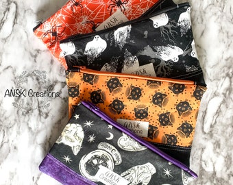 May include: Four Halloween-themed zippered pouches with different patterns. The pouches are stacked on top of each other. The top pouch is orange with black spiders and webs. The second pouch is black with white ghosts and trees. The third pouch is orange with black cats. The bottom pouch is black with white skulls and hands. Each pouch has a white label with the text "ANSK Creations".