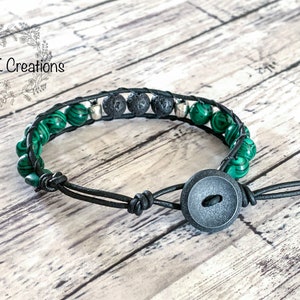 May include: A black leather bracelet with green malachite and black lava beads, silver accents, and a black button closure. ANSK Creations.