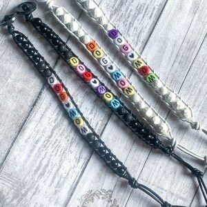 May include: Three bracelets with black and white beads and colorful letter beads spelling out "DOG" and "MOM". The bracelets are on a white wooden surface. ANSK Creations is written in black text on the surface.