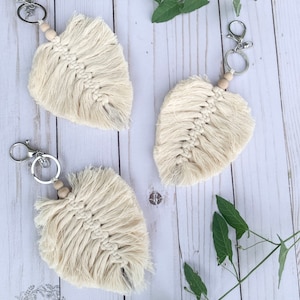 May include: Three cream-colored macrame leaf keychains with wooden beads and silver key rings. The keychains are hanging on a white wooden background with green leafy vines.