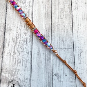 May include: A brown leather bracelet with white and blue beads and a silver charm. The bracelet is tied with a knot at the end.