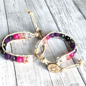 May include: Two leather wrap bracelets with gemstone beads. One bracelet features pink, white, and purple beads. The other bracelet features black, purple, and white beads. Both bracelets have a wooden button closure with a star design. ANSK Creations is printed on the wooden surface.