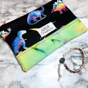May include: A black zippered pencil case with colorful dinosaur illustrations and the text "ANSK Creations". A beaded bracelet with a silver-colored clasp and a beige cord is next to the case.