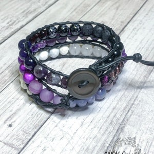 May include: A multi-layered bracelet with purple, white, and black beads wrapped in black leather cord. The bracelet has a black button closure and is accented with a small, black bead.