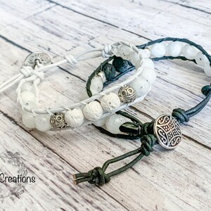 May include: Two white leather bracelets with white and black beads. One bracelet has a silver charm with a geometric design. The bracelets are on a white wooden surface.