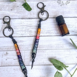 May include: Two macrame keychains with colorful gemstone beads in a rainbow pattern. Each keychain has a black metal clasp and a black cord. A small amber glass bottle with a black cap is also in the image.