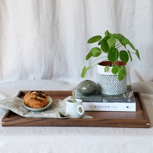 Może przedstawiać: Drewniana taca z białą filiżanką, croissantem i książką zatytułowaną "The Kinfolk Home". Taca znajduje się na białym tle, a w tle znajduje się doniczka z rośliną o okrągłych liściach.