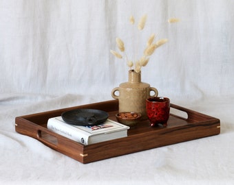 May include: A wooden serving tray with a book, a small bowl of nuts, a ceramic vase with dried flowers, and a red ceramic cup. The tray has two handles on the sides.