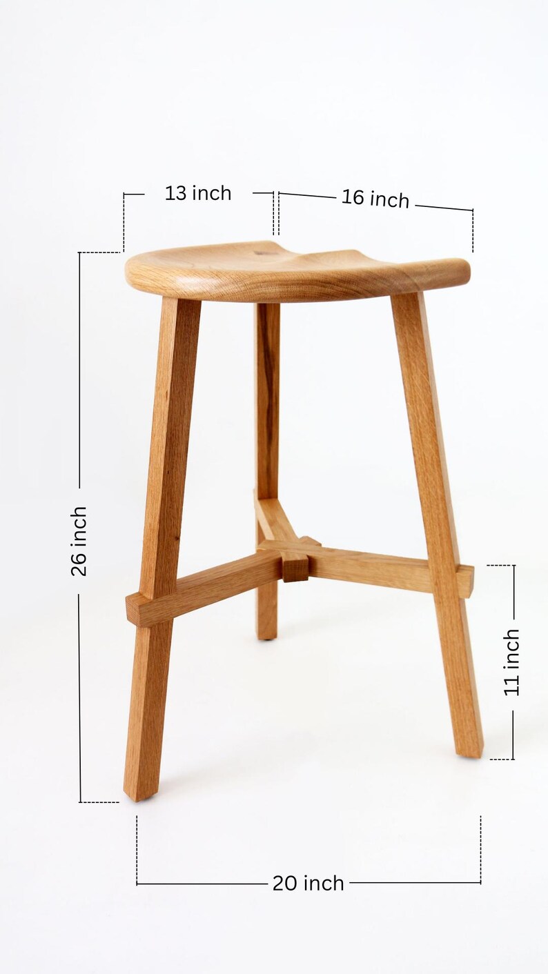 Może przedstawiać: Drewniany stołek z zakrzywionym siedziskiem i trzema nogami. Stołek ma 66 cm wysokości, 41 cm szerokości i 51 cm głębokości.