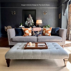 Puede incluir: Un sof&aacute; gris con estampado de patrones con una variedad de almohadas, incluyendo patrones naranja, azul y floral. El sof&aacute; est&aacute; en una sala de estar con una pared gris y una pintura encima. Un otomano gris con botones tapizados se encuentra frente al sof&aacute;.