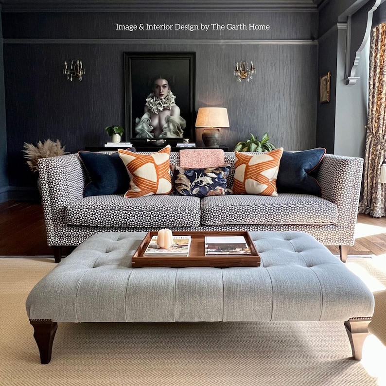 Puede incluir: Un sof&aacute; gris con estampado de patrones con una variedad de almohadas, incluyendo patrones naranja, azul y floral. El sof&aacute; est&aacute; en una sala de estar con una pared gris y una pintura encima. Un otomano gris con botones tapizados se encuentra frente al sof&aacute;.