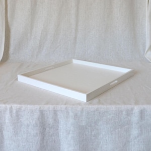 May include: A square, white serving tray with a minimalist design. The tray has a raised edge and cut-out handles on the sides. The tray is made of wood and is painted white. The tray is sitting on a white surface.