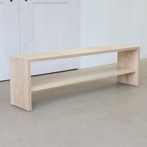 Può includere: Una panca in legno chiaro con uno scaffale sottostante. La panca è realizzata in legno naturale e ha un design semplice e moderno.