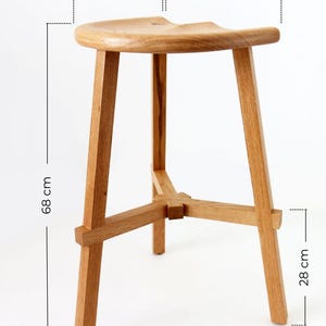 Może przedstawiać: Drewniany stołek z zakrzywionym siedziskiem i trzema nogami. Stołek ma 41 cm szerokości, 33 cm głębokości, 68 cm wysokości i 50 cm średnicy.