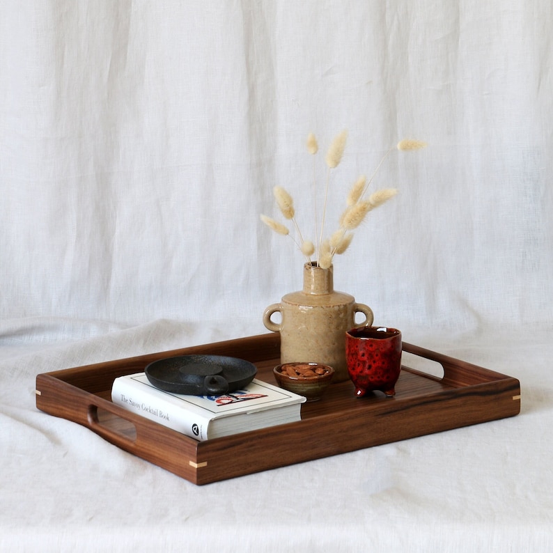 Puede incluir: Una bandeja de servir de madera con un libro, un peque&ntilde;o cuenco de nueces, un jarr&oacute;n de cer&aacute;mica con flores secas y una taza de cer&aacute;mica roja. La bandeja tiene dos asas a los lados.
