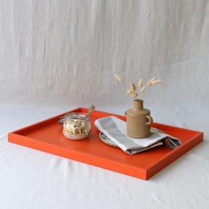 May include: A bright orange rectangular serving tray with a glass jar, a small vase with dried flowers, a plate with a grey and white striped napkin, and a small brown vase. The tray is on a white surface.