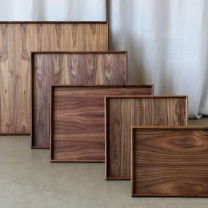 Może przedstawiać: Pięć drewnianych tac drewnianych o różnych rozmiarach, wszystkie wykonane z tego samego ciemnego drewna o bogatym usłojeniu. Tace są ułożone w sposób schodkowy, z największą tacą na górze i najmniejszą tacą na dole.