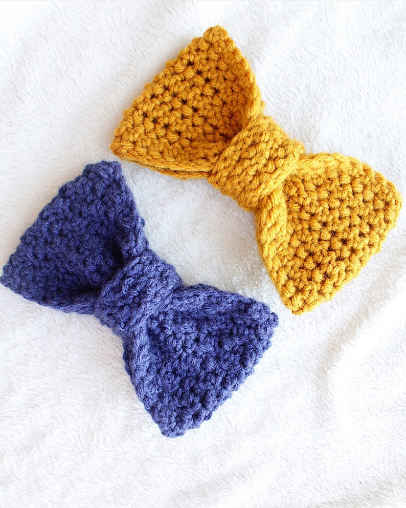 May include: Two crocheted bows, one in a deep blue and one in a mustard yellow. The bows are made with a textured stitch and are tied in a simple knot.