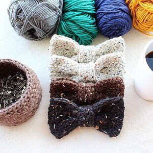 May include: Four crocheted bow ties in different colors and patterns: white with speckles, brown with speckles, black with speckles, and a brown basket filled with safety pins.