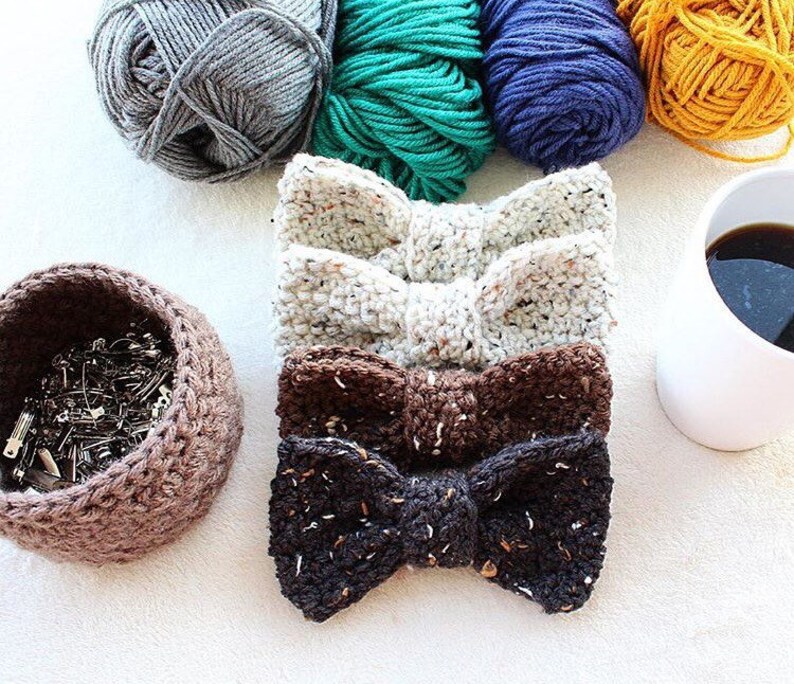 May include: Four crocheted bow ties in different colors and patterns: white with speckles, brown with speckles, black with speckles, and a brown basket filled with safety pins.