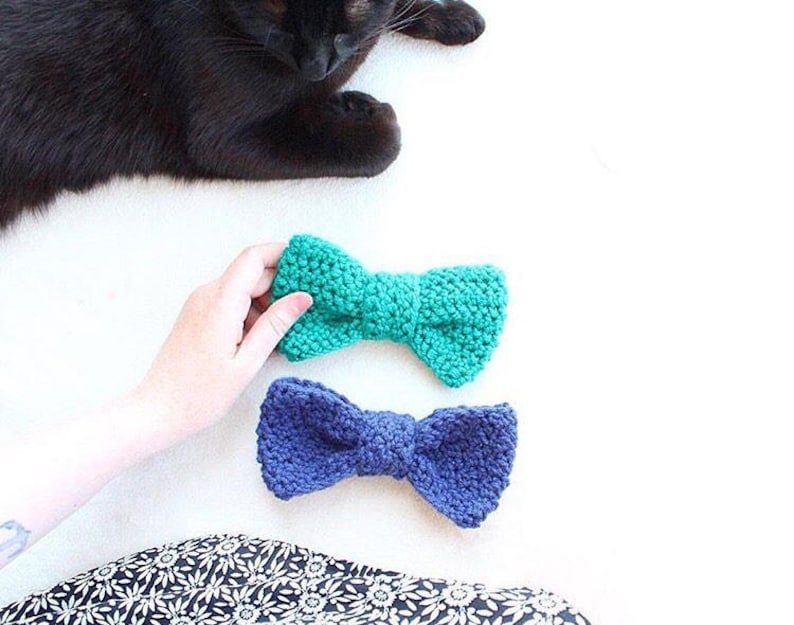 May include: Two crocheted bow ties, one teal and one navy blue, on a white surface.